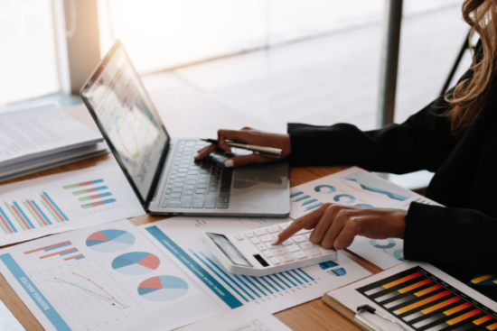 A business professional analyzing financial reports, using a laptop and calculator, with charts and graphs spread across the desk