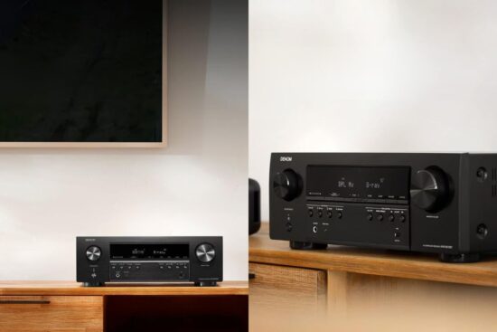 A black Denon AV receiver sits on a wooden shelf under a wall-mounted TV, showing its buttons, knobs, and display screen.