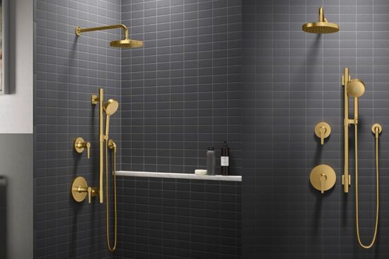 A modern bathroom with dark tiled walls features a gold shower system with a rain showerhead, a hand shower, and matching fixtures.
