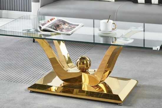A modern coffee table with a glass top, gold metal base, and a curved design, placed on a gray rug in a living room.
