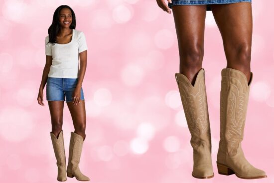 A woman wearing tall beige cowboy boots, denim shorts, and a white top stands against a pink background.