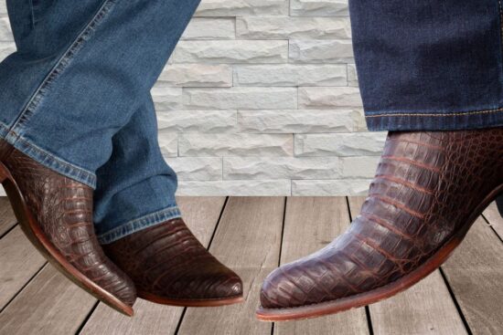 Two pairs of brown caiman leather cowboy boots with pointed toes are worn with blue jeans, standing on a wooden floor against a white brick wall.