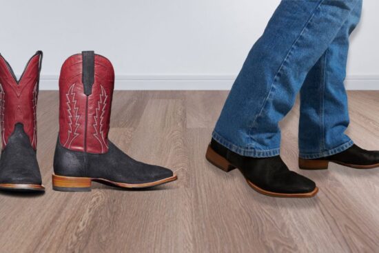 A pair of cowboy boots with black and red leather, one standing upright and the other worn with blue jeans on a wooden floor.
