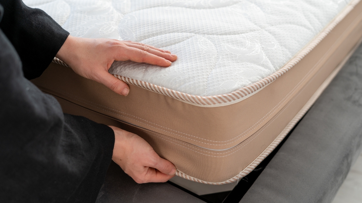 A person in a black sleeve is adjusting a thick, beige and white memory foam mattress on a bed frame.