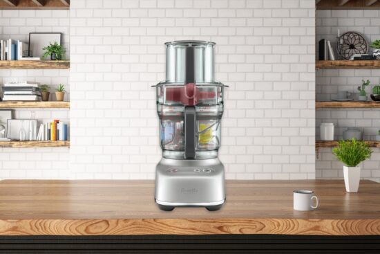 A modern food processor sits on a wooden kitchen counter with shelves and decor in the background.