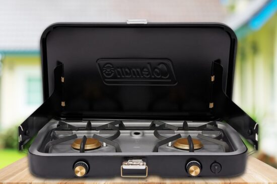 A black Coleman portable gas stove with two burners, set up outdoors on a wooden surface.