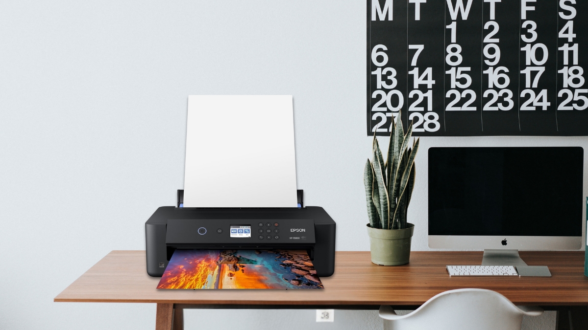 A black Epson printer on a desk printing a colorful photo, next to a computer and a plant.