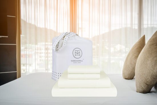 A neatly folded cream-colored bed sheet set with a Threadmill Home Linen bag is placed on a bed with beige pillows in a bright, modern bedroom.
