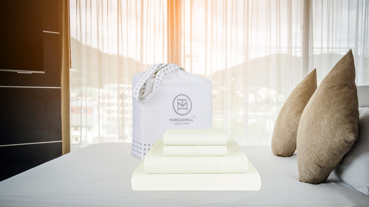 A neatly folded cream-colored bed sheet set with a Threadmill Home Linen bag is placed on a bed with beige pillows in a bright, modern bedroom.