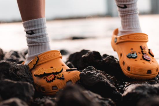 A person wearing orange Crocs with Jibbitz charms, standing on rocky terrain near water at sunset. The Crocs feature the word drew, indicating a collaboration with Drew House.