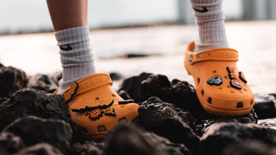 A person wearing orange Crocs with Jibbitz charms, standing on rocky terrain near water at sunset. The Crocs feature the word drew, indicating a collaboration with Drew House.