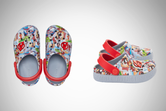 A pair of children's Crocs featuring a colorful Marvel Avengers comic print, with red adjustable heel straps and a grey sole embossed with "Avengers Assemble," displayed against a light grey gradient background.