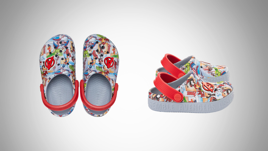 A pair of children's Crocs featuring a colorful Marvel Avengers comic print, with red adjustable heel straps and a grey sole embossed with "Avengers Assemble," displayed against a light grey gradient background.