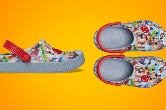 A pair of Marvel-themed Crocs featuring a comic book-style Avengers print, red straps, and "Avengers Assemble" embossed on the grey sole, displayed against an orange background.