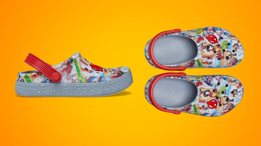 A pair of Marvel-themed Crocs featuring a comic book-style Avengers print, red straps, and "Avengers Assemble" embossed on the grey sole, displayed against an orange background.