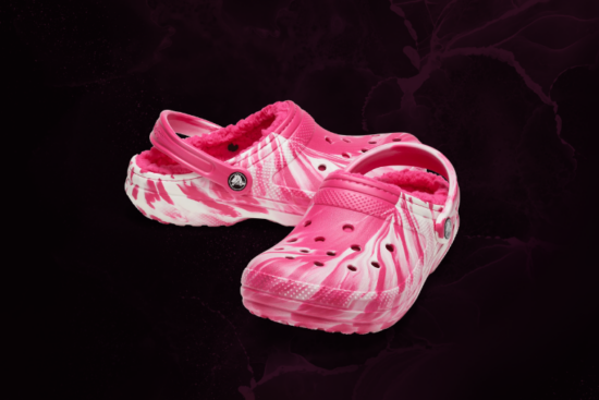 A pair of pink tie-dye Crocs with a plush fuzzy lining, displayed against a dark, textured background.