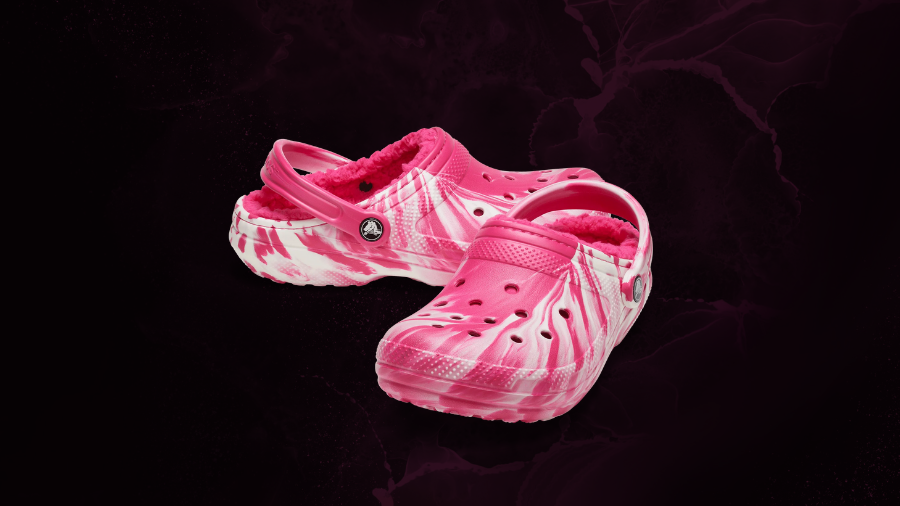 A pair of pink tie-dye Crocs with a plush fuzzy lining, displayed against a dark, textured background.