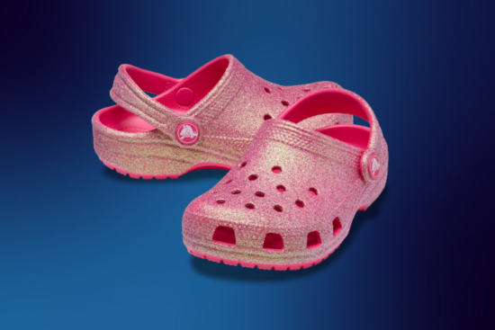 A pair of pink glittery Crocs with a classic clog design, featuring ventilation holes and an adjustable heel strap, displayed against a dark blue gradient background.