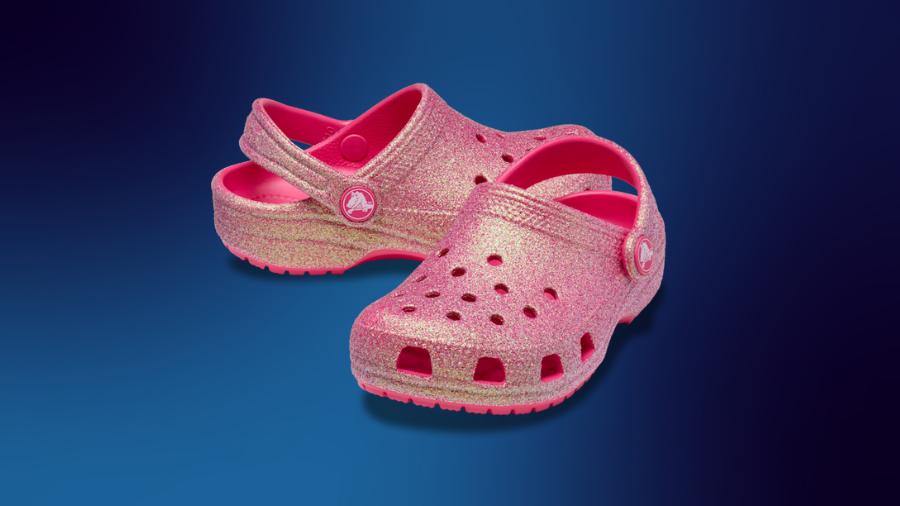 A pair of pink glittery Crocs with a classic clog design, featuring ventilation holes and an adjustable heel strap, displayed against a dark blue gradient background.
