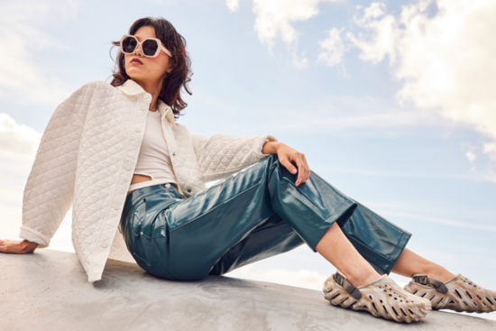 A fashionable woman in a white quilted jacket and blue leather pants, sitting on a ledge wearing tan-colored Crocs with a unique sculpted sole.