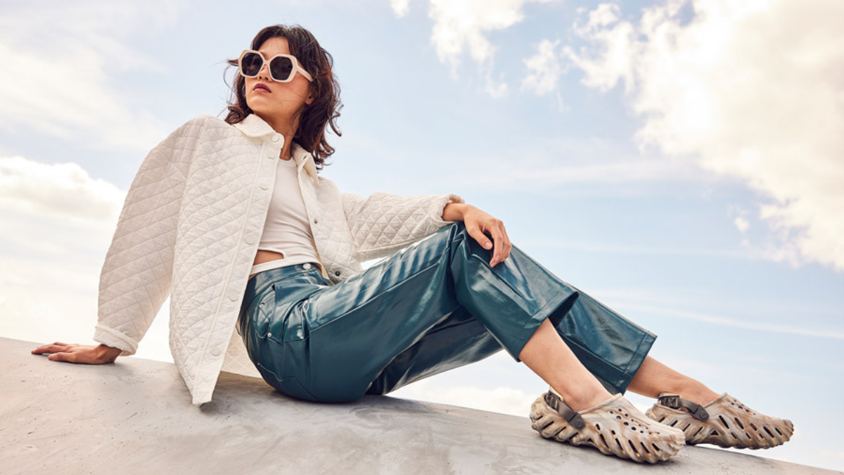 A fashionable woman in a white quilted jacket and blue leather pants, sitting on a ledge wearing tan-colored Crocs with a unique sculpted sole.