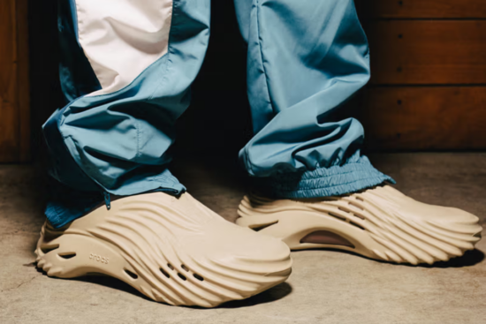 Beige Crocs with a distinctive wave pattern being worn with blue joggers, creating a modern and casual streetwear look.