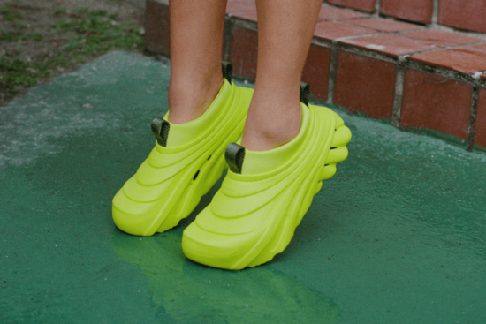 A close-up of a pair of vibrant neon green Crocs with a sculpted, ridged design worn on a green pavement.