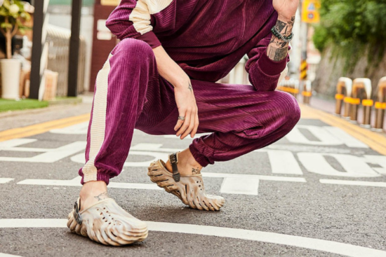 A person squatting on the street wearing a stylish maroon tracksuit and futuristic Crocs with a wavy sole design.