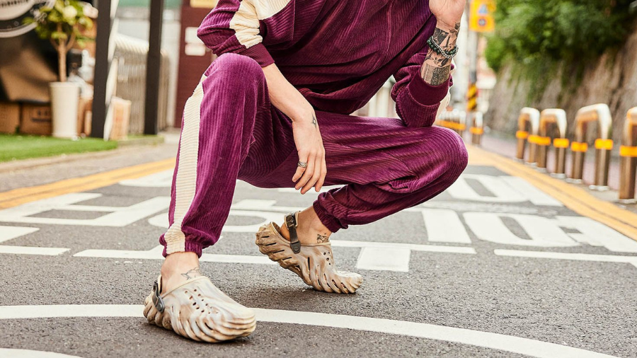A person squatting on the street wearing a stylish maroon tracksuit and futuristic Crocs with a wavy sole design.