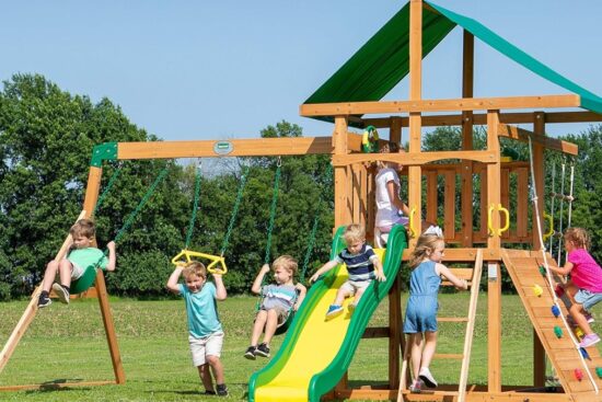 Children playing on the Backyard Discovery Mount McKinley Swing Set, featuring a slide, rock wall, rope ladder, and canopy roof in a grassy backyard.