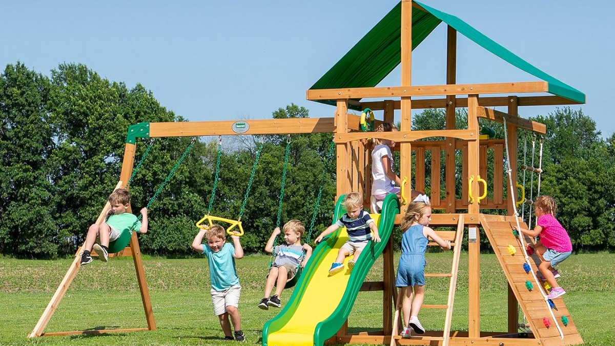 Children playing on the Backyard Discovery Mount McKinley Swing Set, featuring a slide, rock wall, rope ladder, and canopy roof in a grassy backyard.