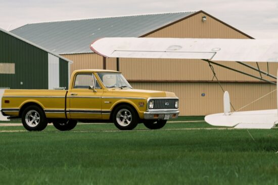 Yellow classic Chevy C10 truck parked on grass beside a vintage airplane near metal buildings.