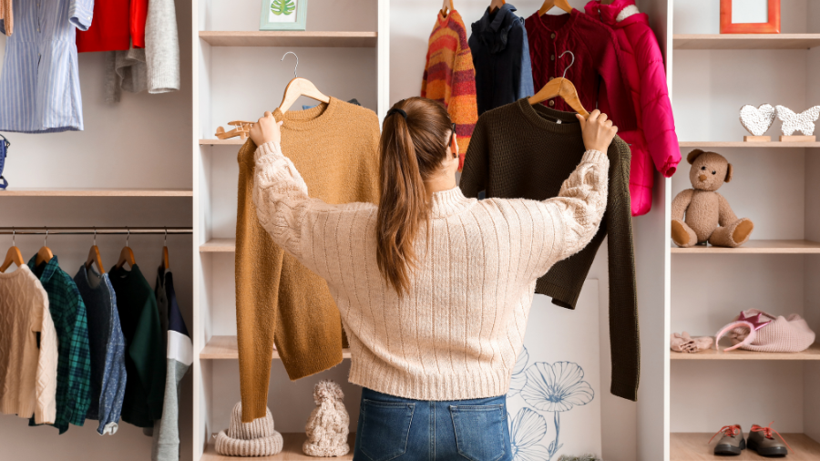 A woman in a cream sweater holding up two knit sweaters, deciding between them in a cozy clothing store.