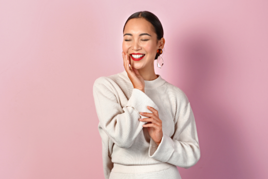 A smiling woman in a light beige ribbed sweater posing with eyes closed against a pink background.