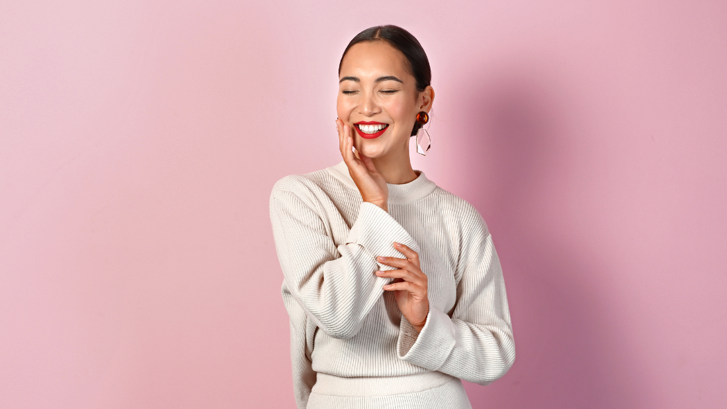 A smiling woman in a light beige ribbed sweater posing with eyes closed against a pink background.