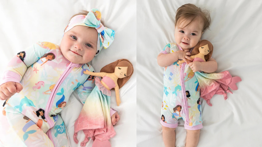 Two babies on a white bed, both wearing pastel pajamas with mermaid prints and each holding or cuddling a soft mermaid doll.