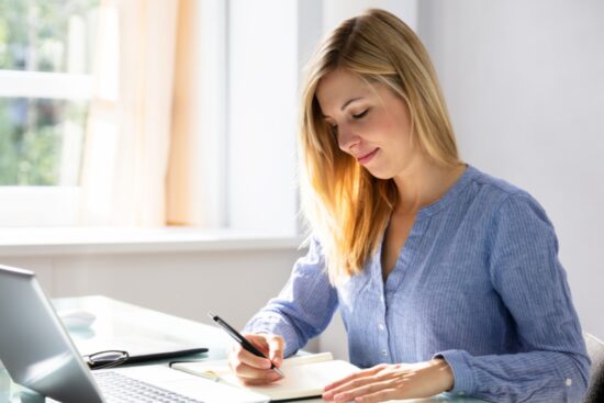 Woman writing in a notebook at her desk with a laptop nearby, using a fountain pen.