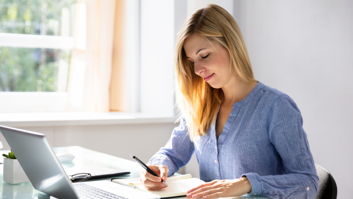 Woman writing in a notebook at her desk with a laptop nearby, using a fountain pen.