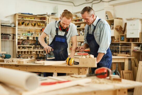 Two men in a woodworking shop using tools and sanding a wooden project at a workbench.