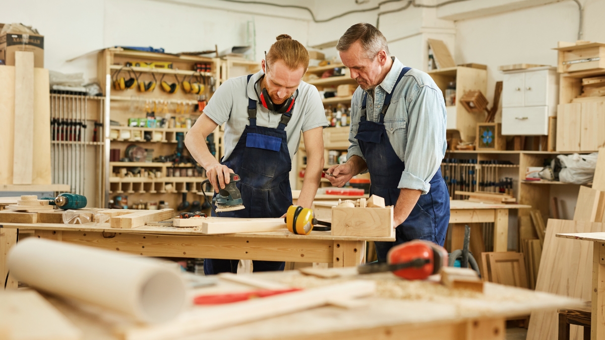 Two men in a woodworking shop using tools and sanding a wooden project at a workbench.