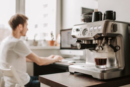 Stainless steel espresso machine brewing coffee in a glass cup on a kitchen counter, with a person working on a computer in the background.