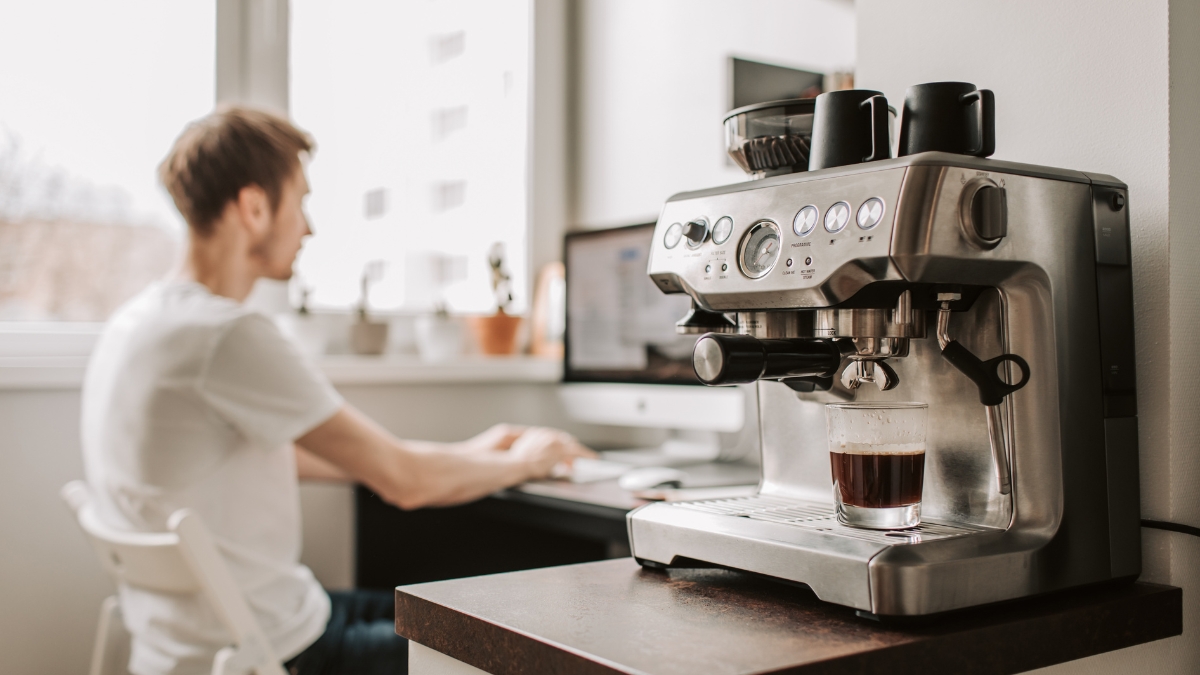 Stainless steel espresso machine brewing coffee in a glass cup on a kitchen counter, with a person working on a computer in the background.