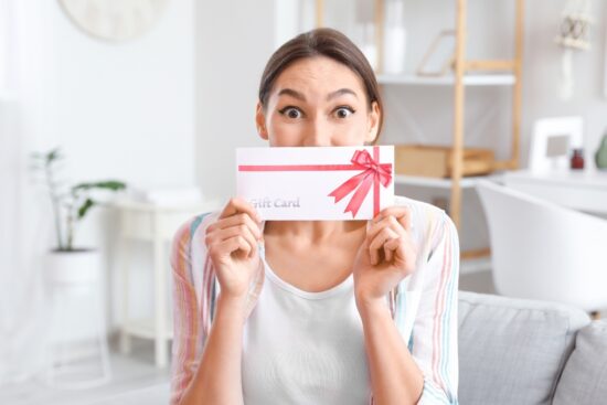 Woman sitting on a couch holding a gift card envelope with a red ribbon and smiling with surprise.
