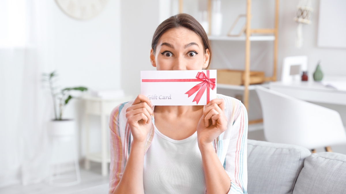 Woman sitting on a couch holding a gift card envelope with a red ribbon and smiling with surprise.