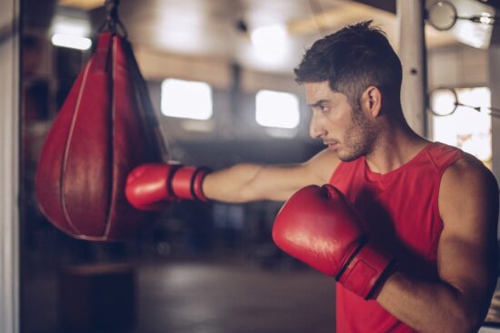 Man wearing red gloves and a sleeveless shirt practicing punches on a red boxing training bag in a gym.