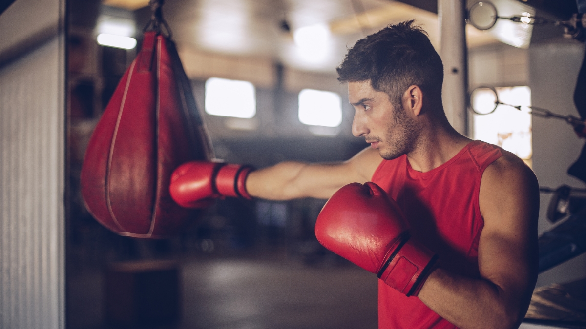 Man wearing red gloves and a sleeveless shirt practicing punches on a red boxing training bag in a gym.