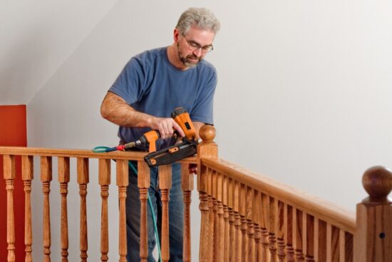 Man using a pneumatic nail gun to install wooden railing on a staircase during a home improvement project.