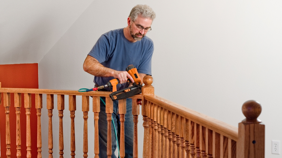 Man using a pneumatic nail gun to install wooden railing on a staircase during a home improvement project.