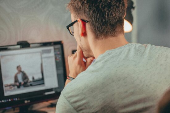 Man wearing glasses editing a photo on a desktop computer in a dimly lit room.