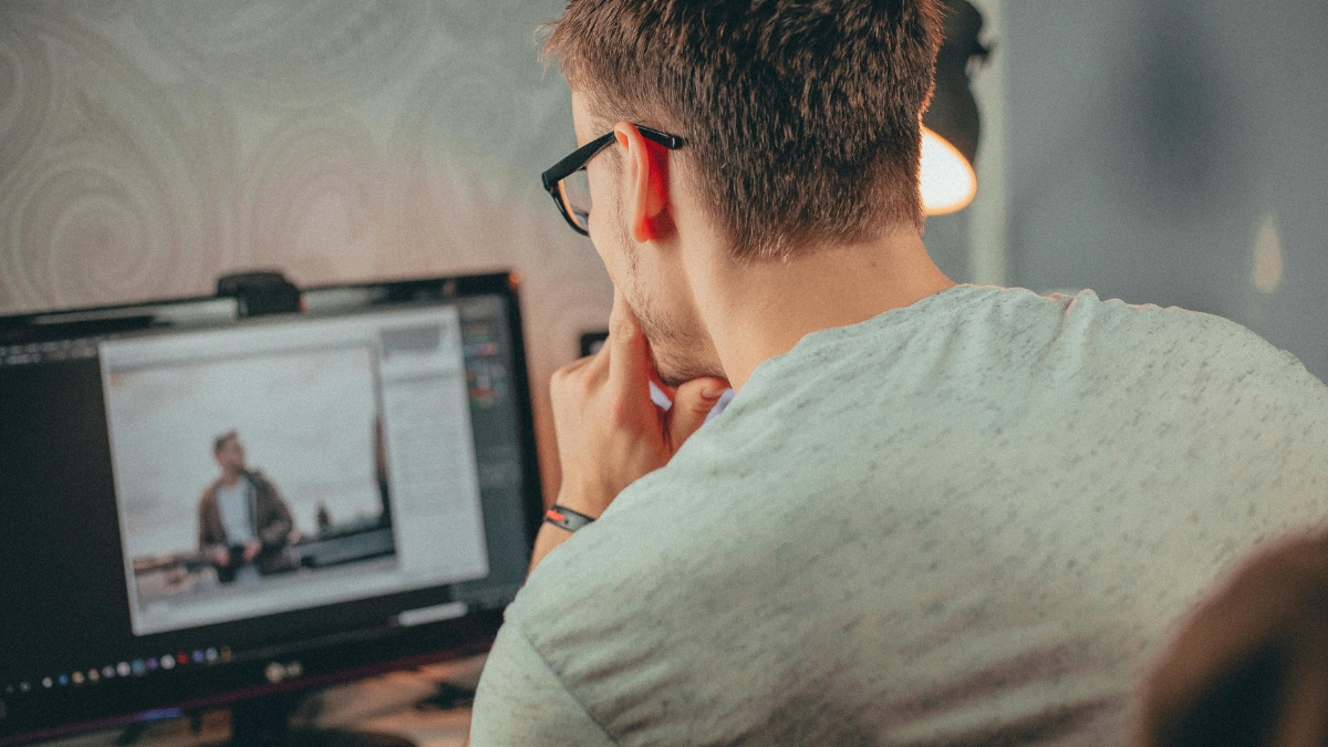 Man wearing glasses editing a photo on a desktop computer in a dimly lit room.